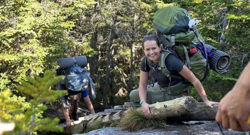 A person wearing a backpack smiles after climbing up on a rock using a ladder. Another person makes their way up behind. 